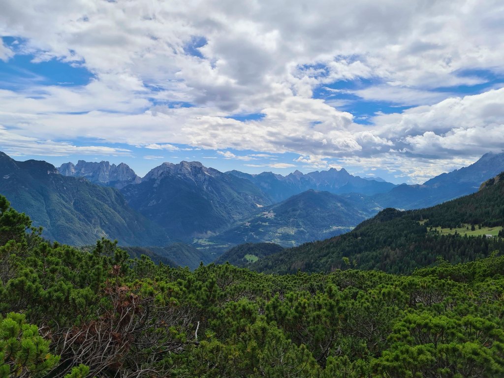 Panorama montano con vette maestose, nuvole e vegetazione lussureggiante in primo piano.