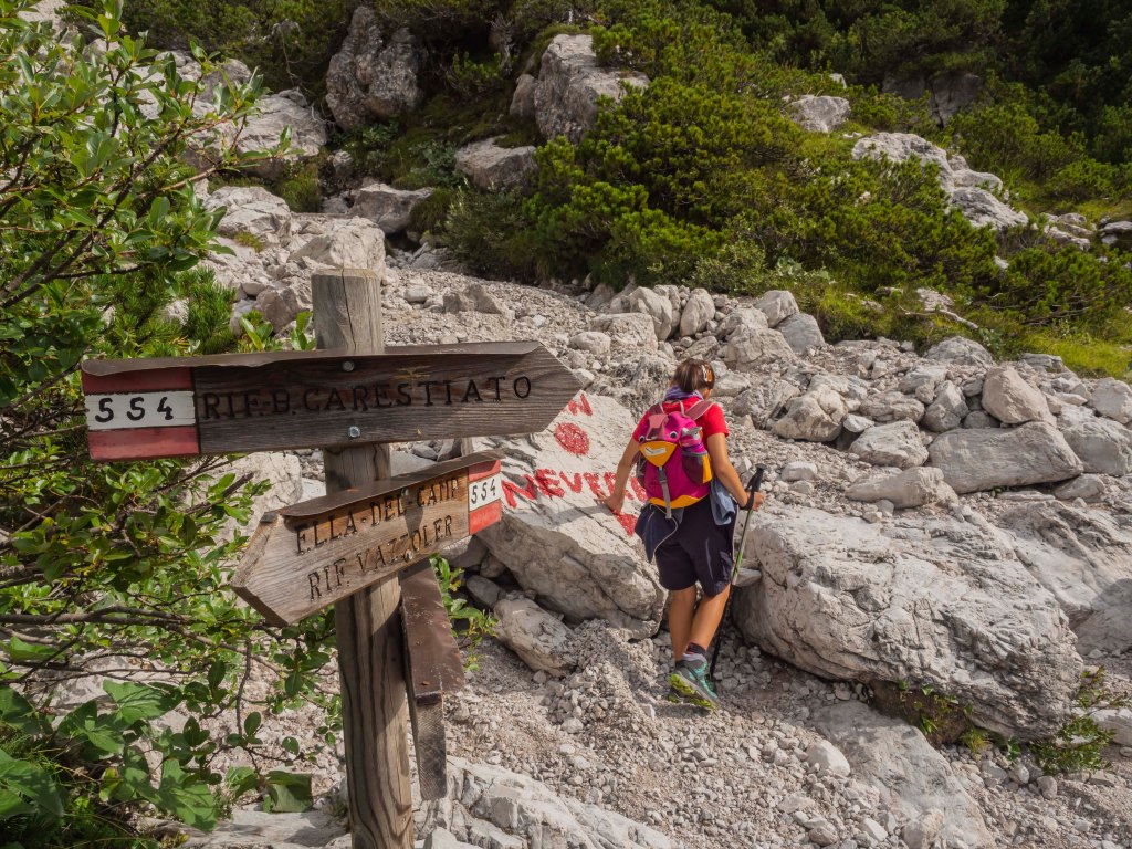 Escursionista che cammina su un sentiero di montagna, con segnaletica legata ai percorsi. Vegetazione verde e rocce nel contesto naturale.