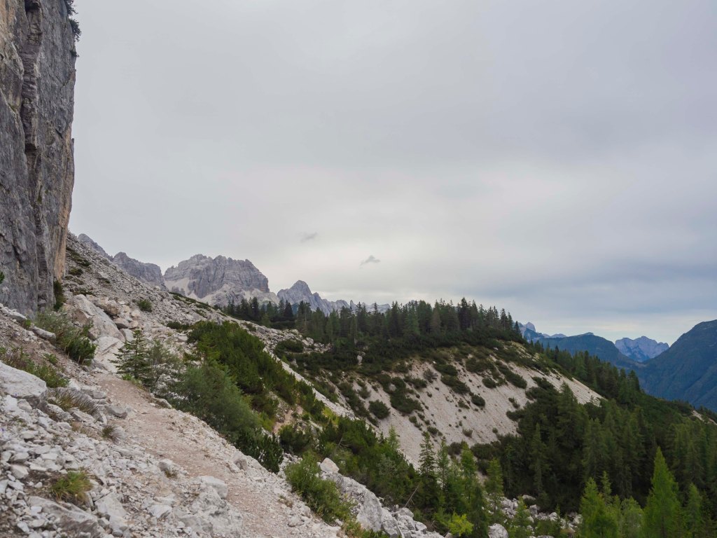 Sentiero montano con vista su boschi e vette alpine sotto un cielo nuvoloso.