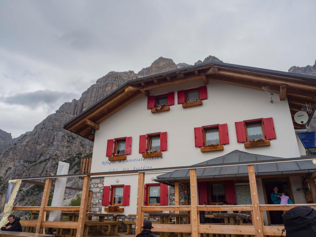 Vista di un rifugio in montagna con finestre con persiane rosse e una terrazza in legno, circondato da montagne. Il cielo è nuvoloso.