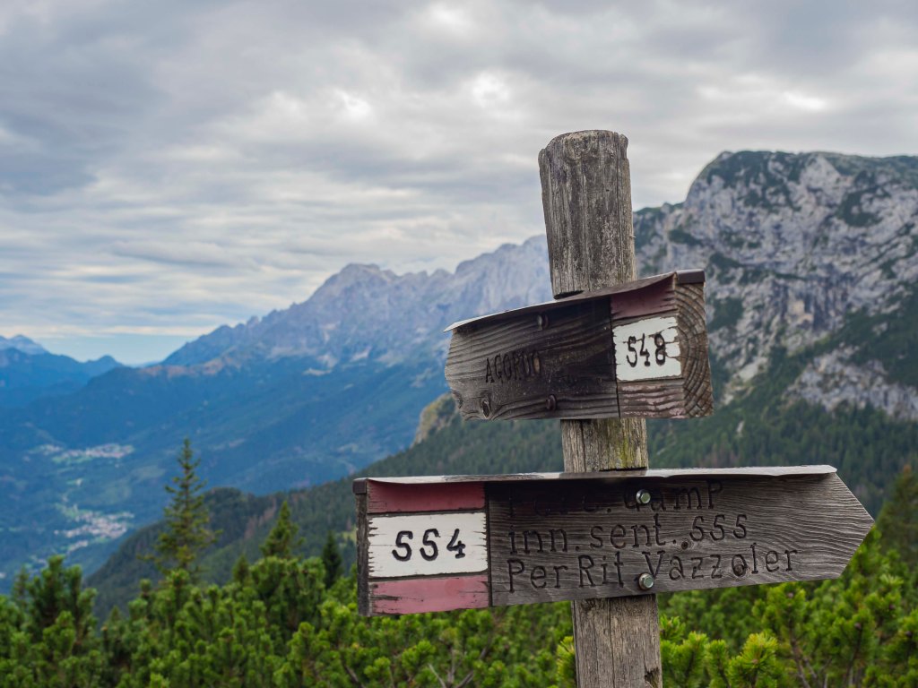 Cartelli stradali in legno indicanti sentieri di montagna, con vista panoramica sulle montagne e nuvole nel cielo.